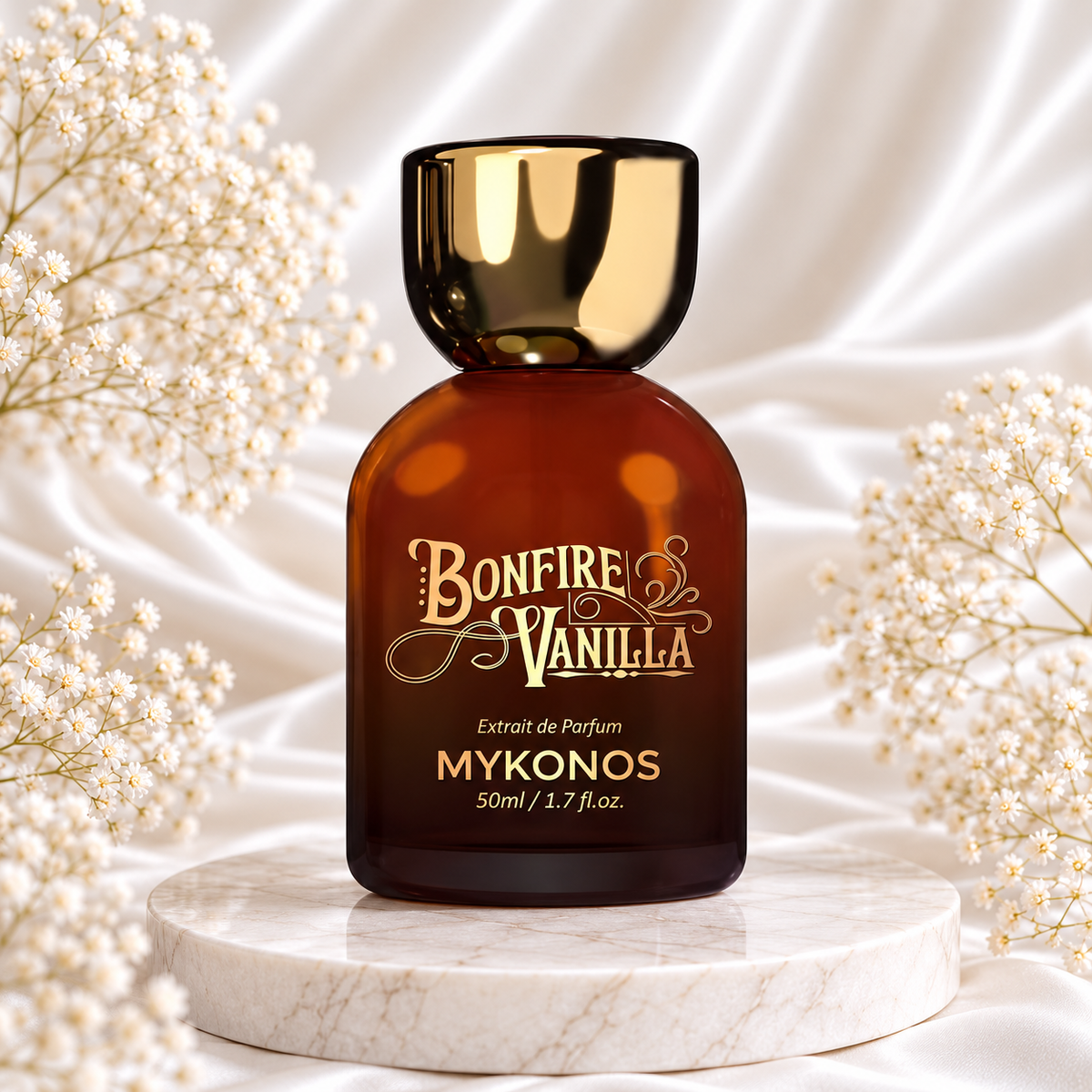 Frasco âmbar de Bonfire Vanilla Extrait de Parfum 50 mL Mykonos, com tampa e inscrições douradas, sobre mármore, entre pequenas flores brancas e tecido acetinado claro.
