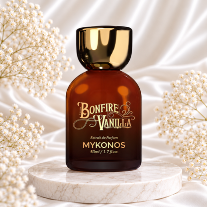 Frasco âmbar de Bonfire Vanilla Extrait de Parfum 50 mL Mykonos, com tampa e inscrições douradas, sobre mármore, entre pequenas flores brancas e tecido acetinado claro.