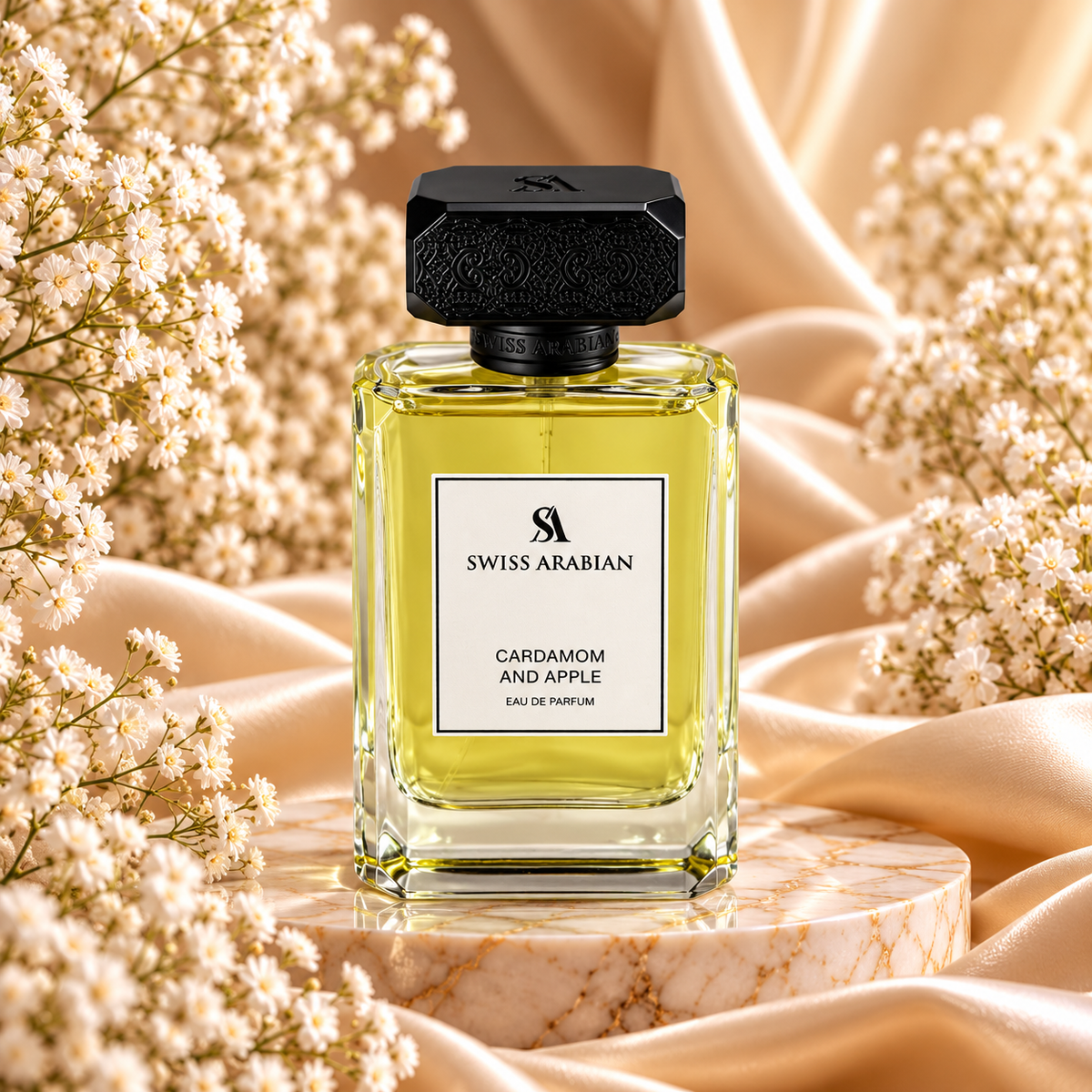 Frasco de Cardamom and Apple Eau de Parfum 100 mL da Swiss Arabian, com líquido verde-amarelado e tampa preta trabalhada, sobre uma base de mármore entre cetim dourado e flores brancas.