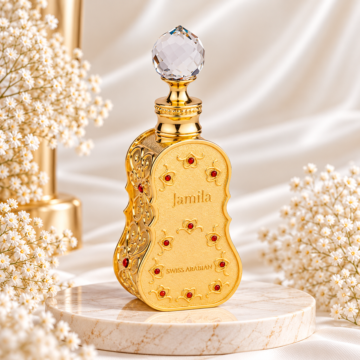 Frasco dourado de Jamila Swiss Arabian, com contornos curvos, decoração trabalhada, pedras vermelhas e tampa transparente facetada, sobre mármore e rodeado de pequenas flores brancas.