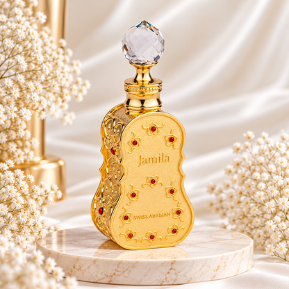 Frasco dourado de Jamila Swiss Arabian, com contornos curvos, decoração trabalhada, pedras vermelhas e tampa transparente facetada, sobre mármore e rodeado de pequenas flores brancas.